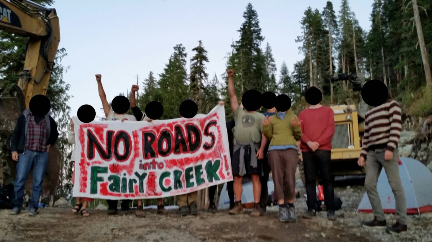 Ten people stand in a row some with their fists raised. They hold a banner saying "No roads into fairy creek". Behind them can be seen heavy machinery and a small grey tent. Further back there are trees.